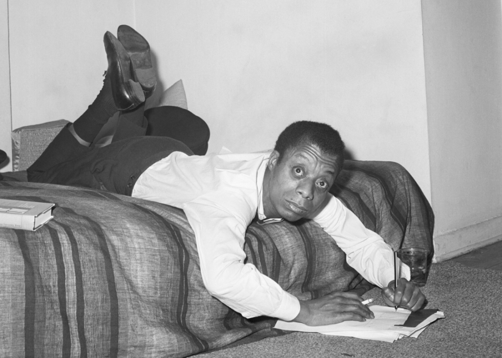 James Baldwin aplecat peste un pat scriind note.